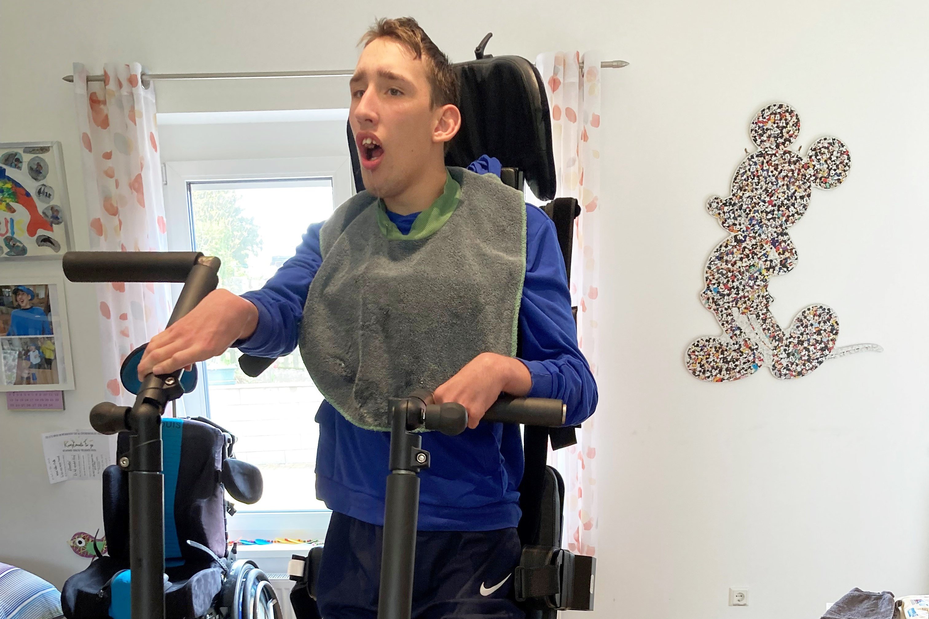 Louis Baumann, ein junger Mann mit Behinderung, steht in einem Stepper. Er legt die H&auml;nde auf die Handgriffe des Ger&auml;ts, und die F&uuml;&szlig;e sind mit Gurten fixiert. Im Hintergrund ist ein Rollstuhl zu sehen. 