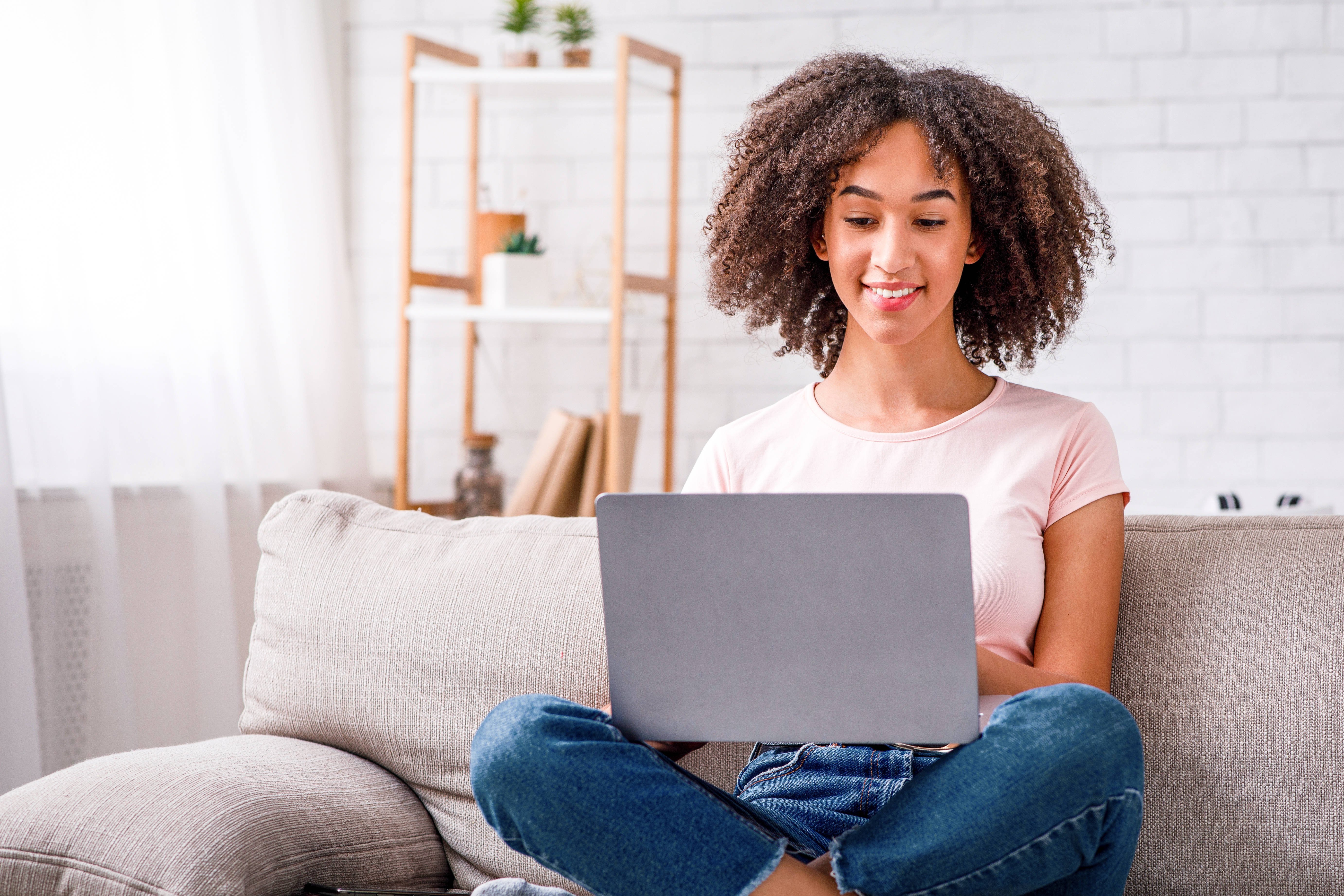 Eine junge Frau sitzt entspannt auf einem Sofa, hat einen Laptop auf dem Scho&szlig; und l&auml;chelt, w&auml;hrend sie auf den Bildschirm schaut. Im Hintergrund steht ein Regal mit Pflanzen und Dekoration.