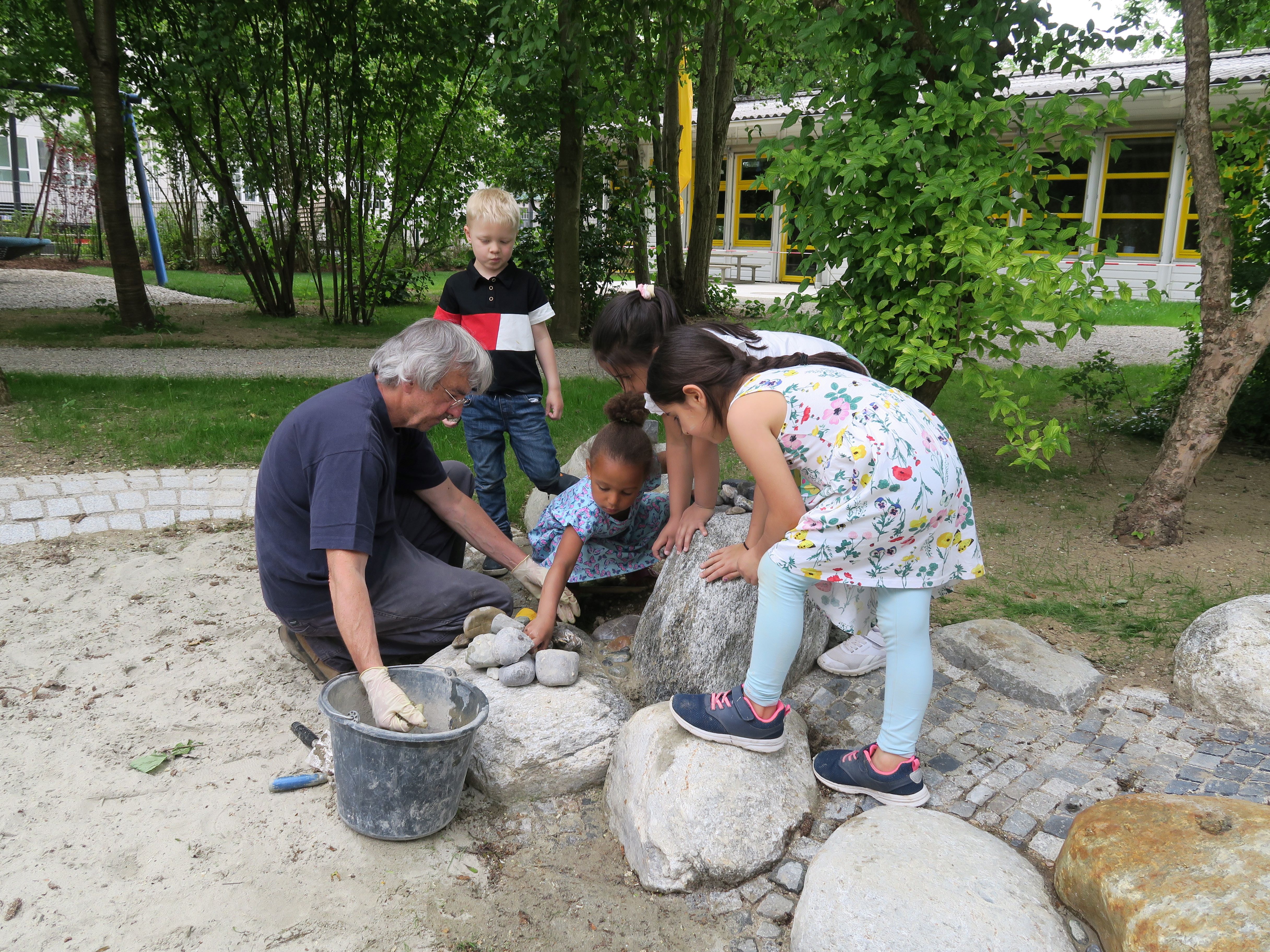 Auf dem Foto sieht man Peter Hohenauer (links), wie er zusammen mit mehreren Kindern einer Münchner Kita-Gruppe ein Mosaik aus mehrfarbigen Steinen gestaltet, die in ein Mörtelbett gesetzt werden.