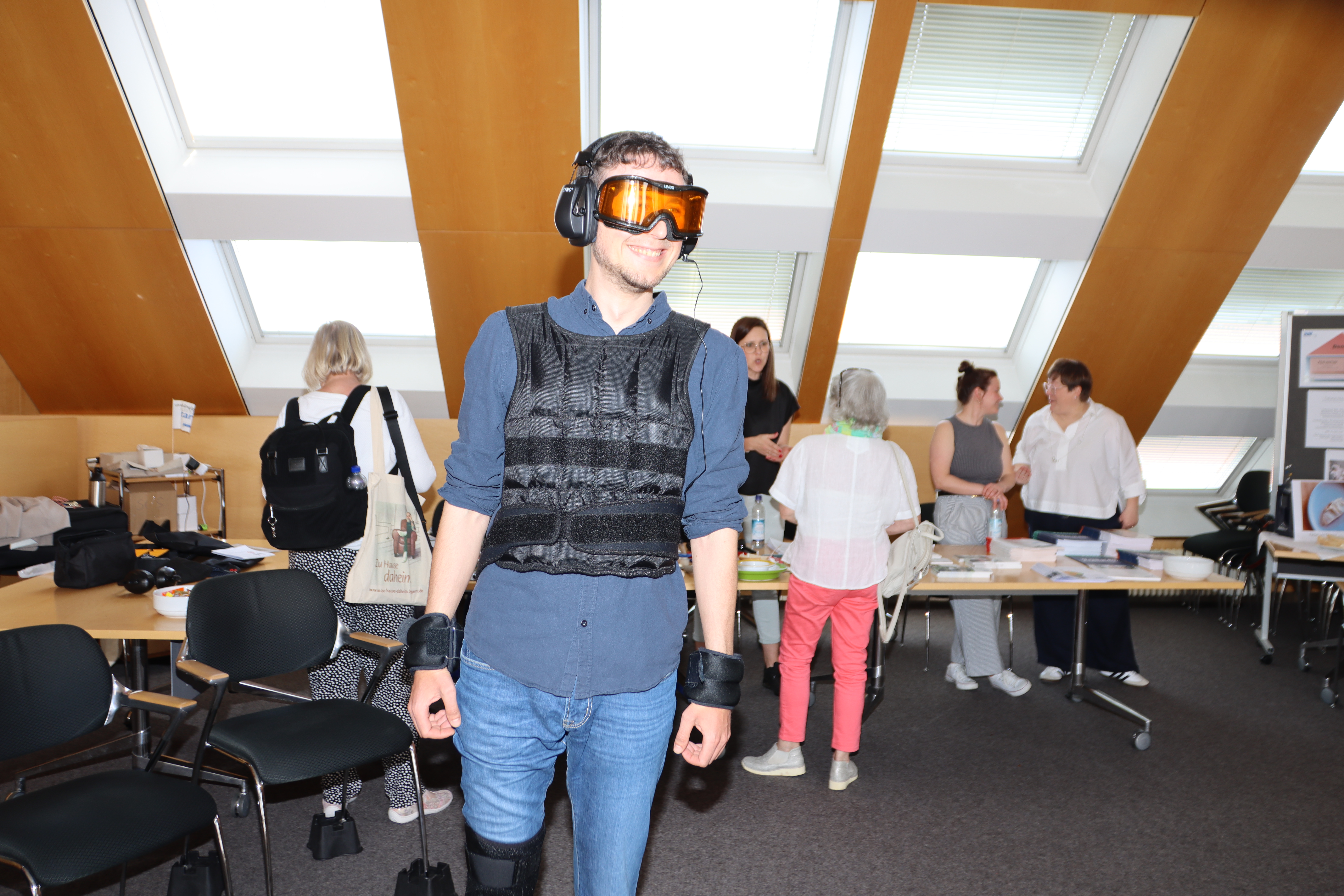 Auf dem Foto sieht man einen Mitarbeitenden des VdK Bayern. Er trägt Kopfhörer, eine orangefarbene Brille, schwarze Bandagen, weiße Handschuhe und eine Weste. Dies soll eingeschränktes Seh- und Hörvermögen simulieren.