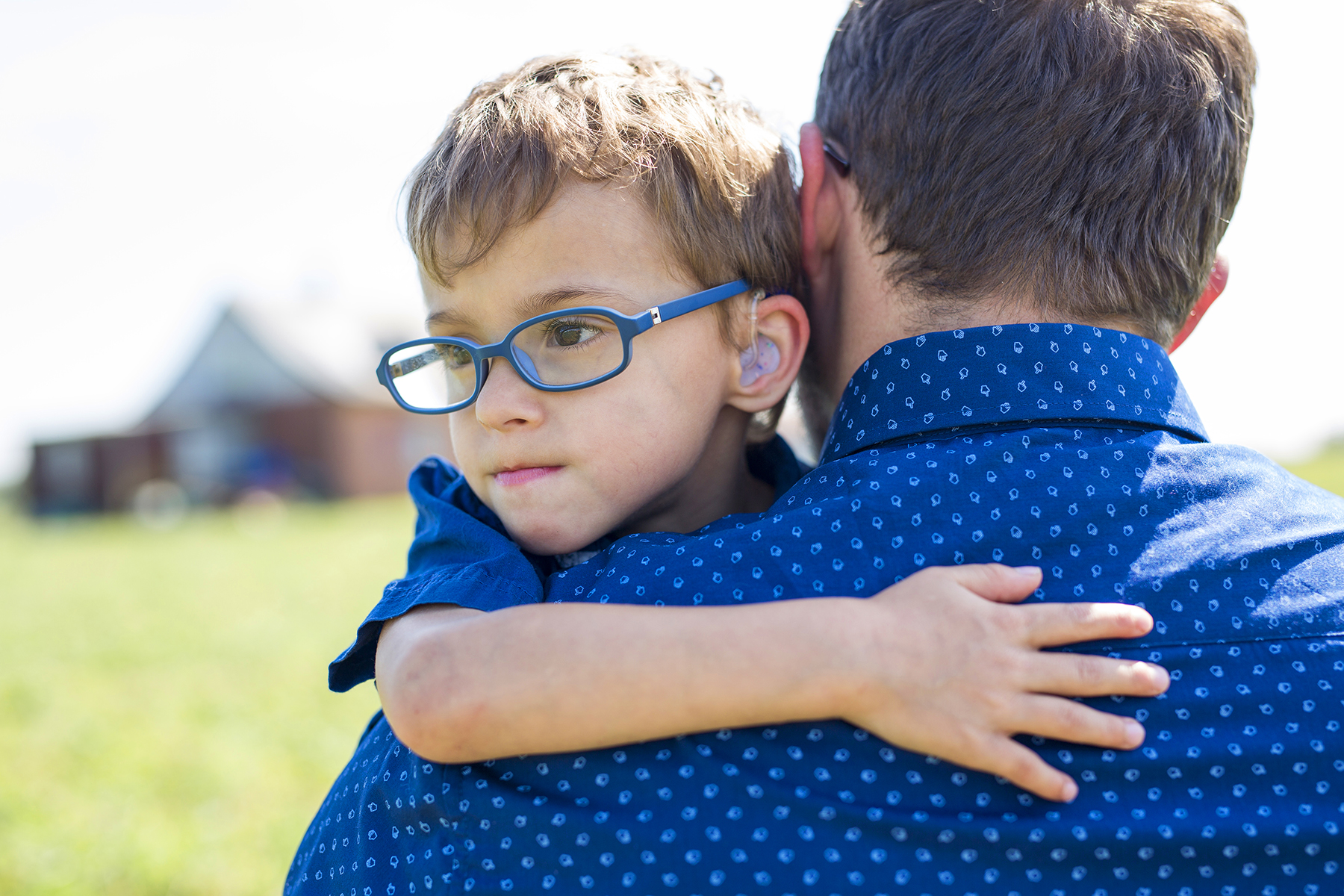 Ein Mann (rechts im Bild) mit dem Rücken zum Fotografen gedreht, hält einen kleinen Jungen (links) auf den Armen. Er trägt eine Brille und ein Hörgerät.