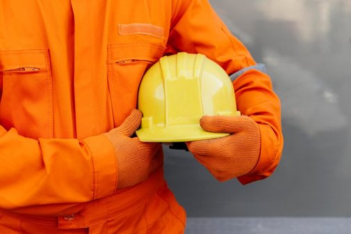 Ein Mensch in einem orangefarbenen Overall trägt einen gelben Helm in den Händen