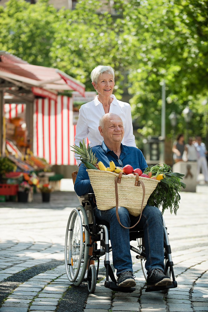 Ein &auml;lteres Paar drau&szlig;en unterwegs, sie schiebt ihn im Rollstuhl. Er hat gef&uuml;llte Einkaufst&uuml;ten auf dem Scho&szlig;.