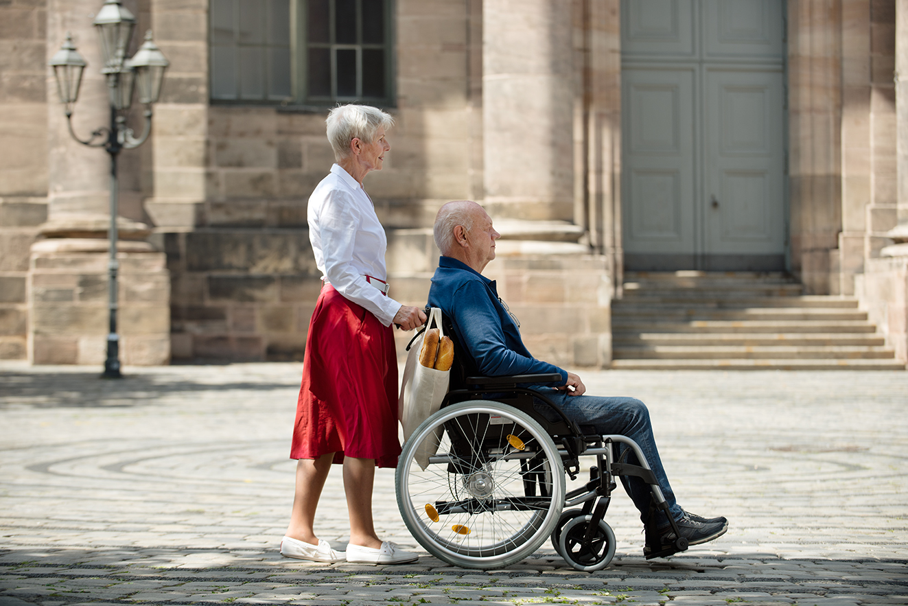 Eine Seniorin schiebt ihren Mann, der im Rollstuhl sitzt, &uuml;ber einen Platz