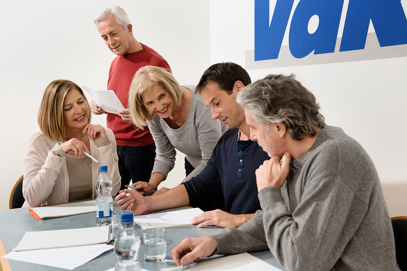 Verschiedene Menschen sitzen an einem Tisch, offenbar in einer Arbeitssitzung, an der Wand hinter ihnen ist ein großes VdK-Logo.