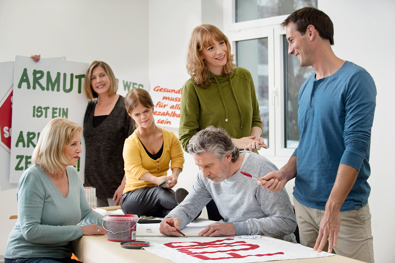 Eine Gruppe unterschiedlicher Menschen - M&auml;nner, Frauen, Alte und Junge, mit und ohne Behinderung, malt gemeinsam Demo-Plakate.