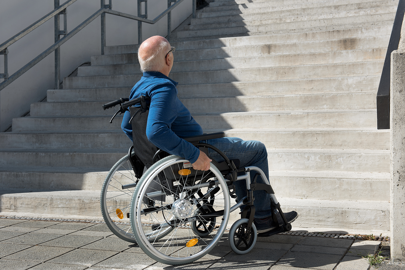 Ein &auml;lterer Mann im Rollstuhl steht ratlos am Fu&szlig; einer hohen Treppe, er schaut die Stufen hinauf. 