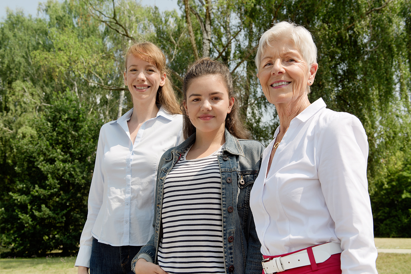 Drei Frauen l&auml;cheln gemeinsam in die Kamera: Eine j&uuml;ngere, eine Teenagerin und eine &auml;ltere Frau.