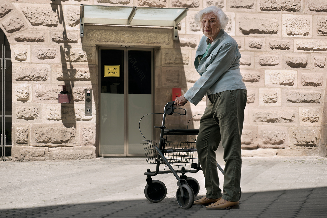 Eine Seniorin mit Rollator steht vor einem Aufzug mit dem Hinweis "Au&szlig;er Betrieb". Sie blickt mit kritischem Blick in die Kamera.