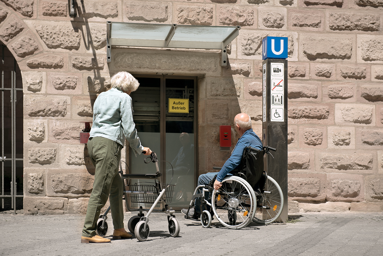 Eine Seniorin mit Rollator und ein Senior im Rollstuhl stehen am unzug&auml;nglichen Eingang einer U-Bahn-Station. Dort ist ein Aufzug mit dem Hinweis "Au&szlig;er Betrieb". 