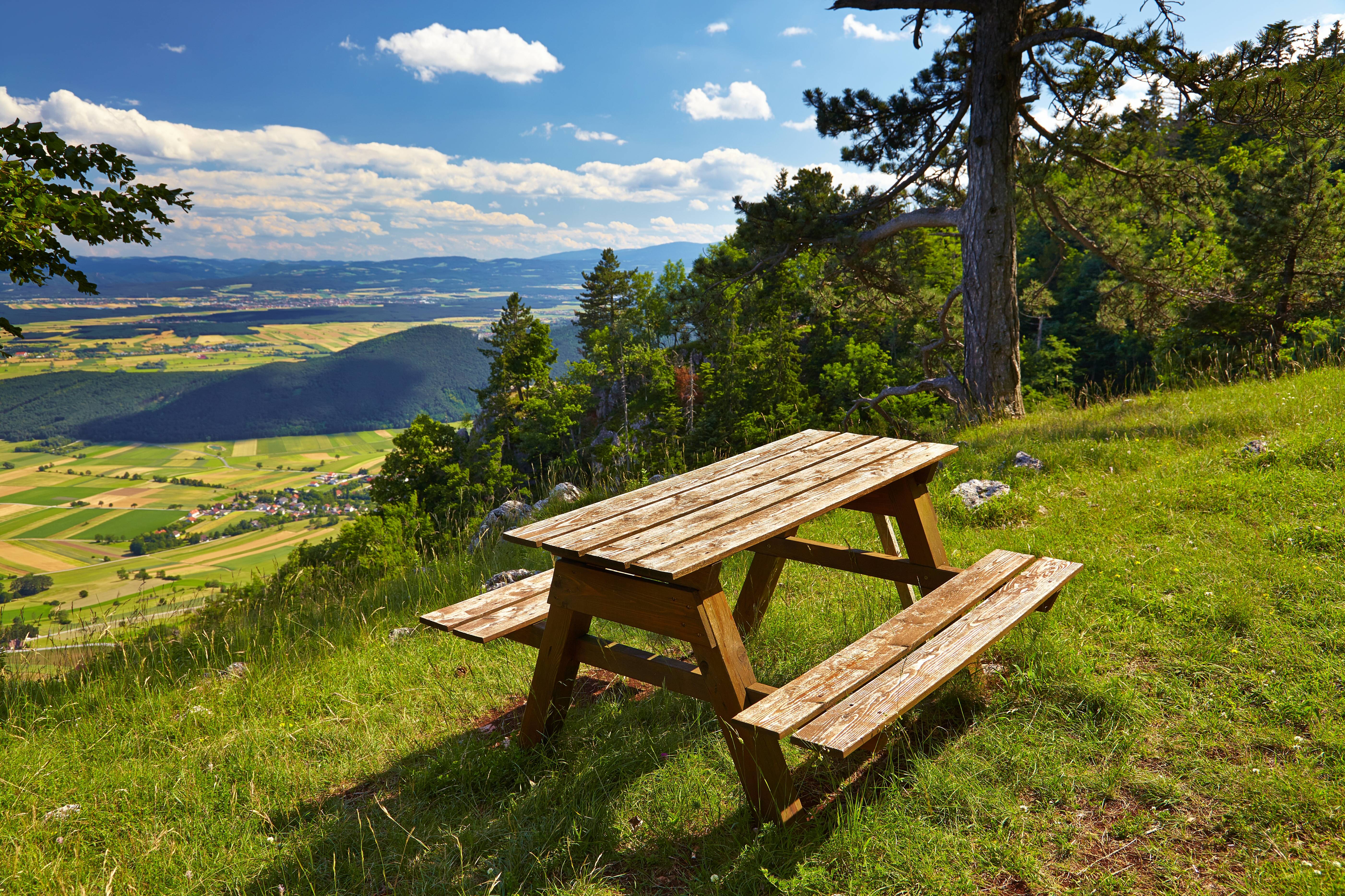 Eine Holzbank mit Tisch steht auf einer Wiese. Im Hintergrund sieht man eine weitl&auml;ufige Landschaft. 