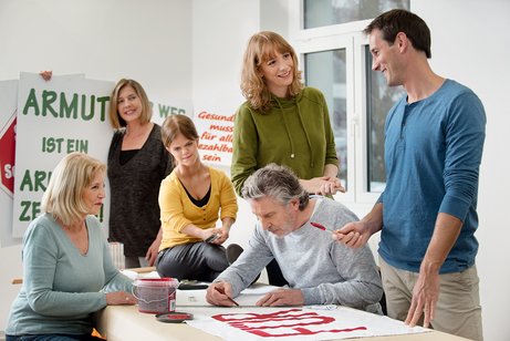 Besprechung Eine Gruppe unterschiedlicher Menschen - Männer, Frauen, Alte und Junge, mit und ohne Behinderung, malt gemeinsam Demo-Plakate.