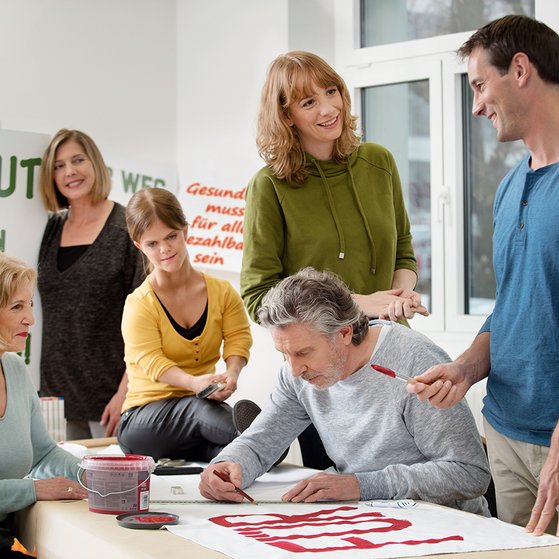 Eine Gruppe unterschiedlicher Menschen - M&auml;nner, Frauen, Alte und Junge, mit und ohne Behinderung, malt gemeinsam Demo-Plakate.
