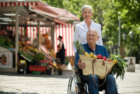 Ein &auml;lteres Paar drau&szlig;en unterwegs, sie schiebt ihn im Rollstuhl. Er hat gef&uuml;llte Einkaufst&uuml;ten auf dem Scho&szlig;.