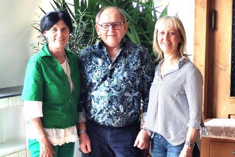 Auf dem Bild stehen drei l&auml;chelnde Erwachsene nebeneinander in einem Raum. Im Hintergrund befinden sich gro&szlig;e gr&uuml;ne Zimmerpflanzen. Die Person links ist die neue erste Vorsitzende, sie tr&auml;gt ein gr&uuml;nes Outfit mit wei&szlig;en Schuhen. Die Person in der Mitte ist der zweite Vorsitzende, er tr&auml;gt ein dunkel gemustertes Hemd, dunkle Hose und Brille. Rechts steht eine Person, die neue Beauftragte der Frauen, mit blondem Haar in einer gestreiften Bluse, Jeans und wei&szlig;en Sneakers.
