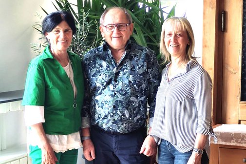 Auf dem Bild stehen drei l&auml;chelnde Erwachsene nebeneinander in einem Raum. Im Hintergrund befinden sich gro&szlig;e gr&uuml;ne Zimmerpflanzen. Die Person links ist die neue erste Vorsitzende, sie tr&auml;gt ein gr&uuml;nes Outfit mit wei&szlig;en Schuhen. Die Person in der Mitte ist der zweite Vorsitzende, er tr&auml;gt ein dunkel gemustertes Hemd, dunkle Hose und Brille. Rechts steht eine Person, die neue Beauftragte der Frauen, mit blondem Haar in einer gestreiften Bluse, Jeans und wei&szlig;en Sneakers.