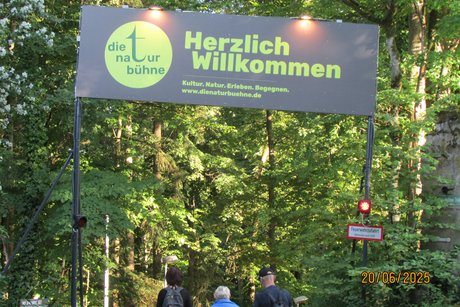 Waldweg mit einem gro&szlig;en Schild auf dem steht Herzlich Willkommen zur Naturb&uuml;hne Trebgast und Menschen die unter dem Schild laufen