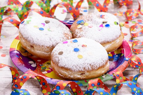 Drei mit Puderzucker best&auml;ubte Krapfen liegen auf einem bunten Pappteller, umgeben von farbenfrohem Konfetti und gemusterten Luftschlangen auf einem Holztisch.