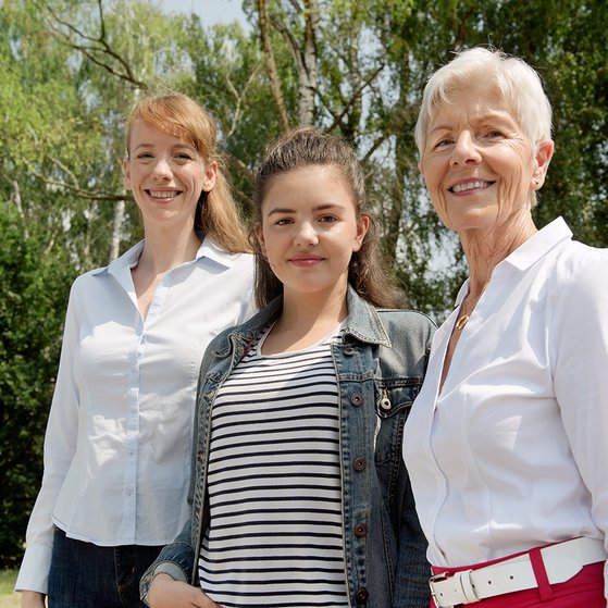 Drei Frauen l&auml;cheln gemeinsam in die Kamera: Eine j&uuml;ngere, eine Teenagerin und eine &auml;ltere Frau.