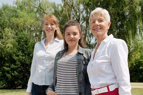 Drei Frauen l&auml;cheln gemeinsam in die Kamera: Eine j&uuml;ngere, eine Teenagerin und eine &auml;ltere Frau.