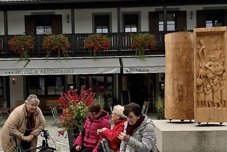 Bild von warten 4 Ausflugsteilnehmer, die auf den Bus vorm "s'Wirtshaus Oberammergau" warten, im Hintergrund der "Kofel"