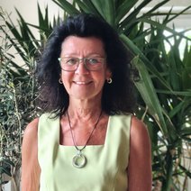 Frau Reck, Kassiererin, l&auml;chelt freundlich, tr&auml;gt eine Brille, hat lange dunkle Haare tr&auml;gt ein hellgr&uuml;nes Oberteil und eine Kette mit einem runden Anh&auml;nger.