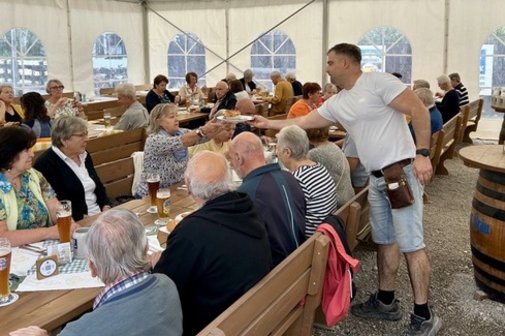 in einem kleinen Bierzelt serviert die Bedienung Wei&szlig;w&uuml;rste, Menschen sitzen gesellig an Biertischen