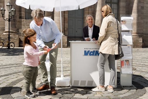 VdK-Infostand Ein VdK-Infostand mit einer Mitarbeiterin, davor verschiedene Menschen, die sich informieren wollen.