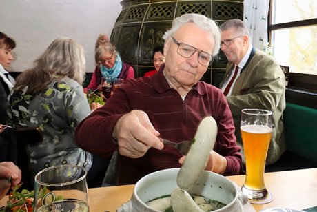 Die Teilnehmer lassen sich die Weißwürste schmecken Das Bild zeigt einen Teilnehmer, der sich ein Paar Weißwürste aus der Terrine ist. Im Hintergrund sind weitere Gäste zu sehen.