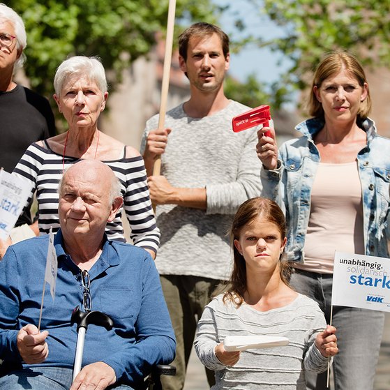 Gruppe von Menschen verschiedenen Geschlechts und Alters, mit und ohne Behinderung, demonstrieren. Auf einer Demopappe steht "Pflege darf nicht arm machen"