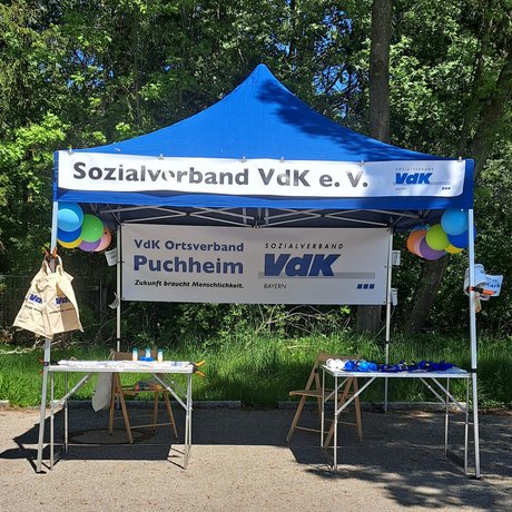 Ein blauer Pavillon durch weiße Banner als Infostand des VdK Puchheim kenntlich gemacht. Er ist mit Luftballons und Fähnchen geschmückt. Darunter sind 2 Tische mit Info- und Werbematerial, sowie zwei leere Klappstühle aufgestellt.