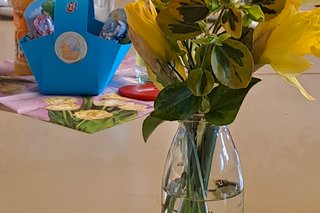 Auf dem Bild sieht man einen Tisch eine Vase mit einem Strauß Osterglocken, dahinter ein Osterkörbchen in blau und Getränke.