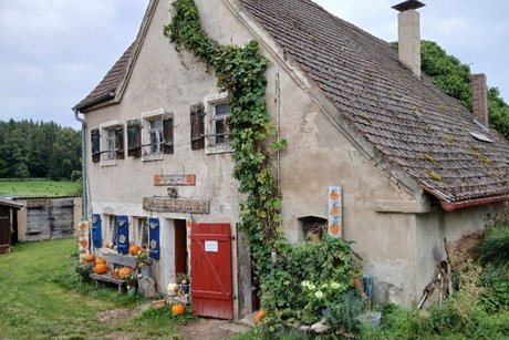 kleines altes Ausstellungshaus auf dem Kürbishof Die Vorderfront eines sehr alten kleines Häuschens, dass mit kleinen Zimmer und immer andere alte Gegenstände mit Kürbissen bestückt ist. Vor dem Haus eine alte braune Haustüre und eine Gartenbank, belegt mit Kürbissen. Eine Blumenranke wächst bis zum Dach.