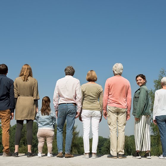 Menschen verschiedenen Alters mit und ohne Behinderung stehen nebeneinander, wenden dem Betrachter den Rücken zu. Eine Teenagerin blickt sich um, sieht den Betrachter direkt an.