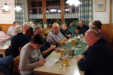 Mitglieder sitzen in der Gaststube mit Getr&auml;nken an einem langen Tisch. Im Vordergrund schreibt eine Frau eine Gru&szlig;karte.