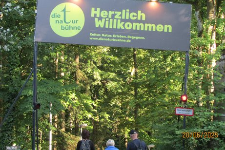 Waldweg mit einem großen Schild auf dem steht Herzlich Willkommen zur Naturbühne Trebgast und Menschen die unter dem Schild laufen