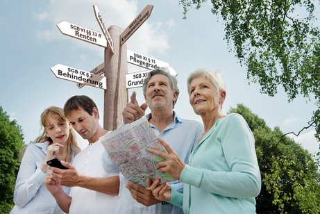 Ein j&uuml;ngeres und ein &auml;lteres P&auml;rchen suchen mit einer Karte nach etwas, hinter ihnen ein Wegweiser, auf dem verschiedene Begriffe aus dem Sozialrecht stehen. 