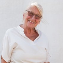 Ingrid Steger, Beisitzerin und kommissarische Schriftführerin des VdK Ortsverbands Aubing, steht vor einem hellen Hintergrund und lächelt freundlich in die Kamera. Sie trägt eine weiße Bluse und eine auffällig gemusterte Hose mit buntem Blumenmotiv. Neben ihr sitzt ihre Hündin Ema, ein großer, langhaariger schwarzer Hund mit braunen Abzeichen, der aufmerksam zur Seite schaut.