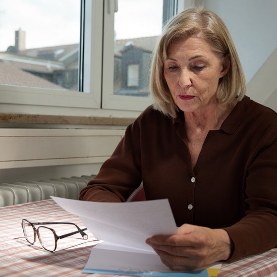 Eine &auml;ltere Frau sitzt an einem K&uuml;chentisch und liest einen Brief, sie sieht ernst und besorgt aus. 