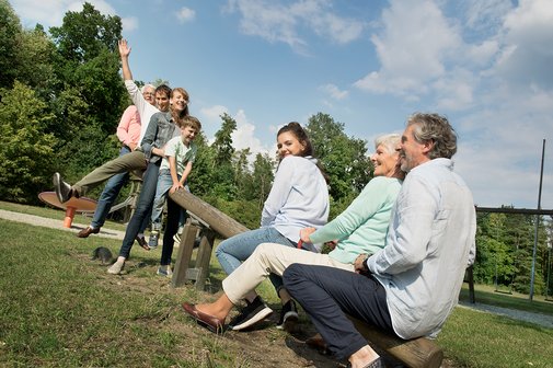 Junge und alte Menschen und ein Kind sitzen auf einer Wippe auf einem Spielplatz Junge und alte Menschen und ein Kind sitzen auf einer Wippe auf einem Spielplatz