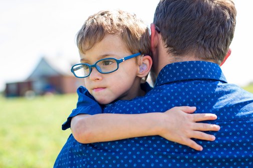 Ein kleiner Junge mit blauer Brille und H&ouml;rger&auml;ten auf dem Arm seines Vaters