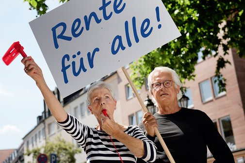 Eine &auml;ltere Frau und ein &auml;lterer Mann mit einer Demopappe mit der Aufschrift "Rente f&uuml;r alle!". Die Frau bl&auml;st in eine Trillerpfeife, h&auml;lt eine Ratsche in der Hand. 