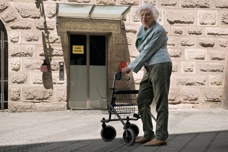 Eine Seniorin mit Rollator steht vor einem Aufzug mit dem Hinweis "Au&szlig;er Betrieb". Sie blickt mit kritischem Blick in die Kamera.