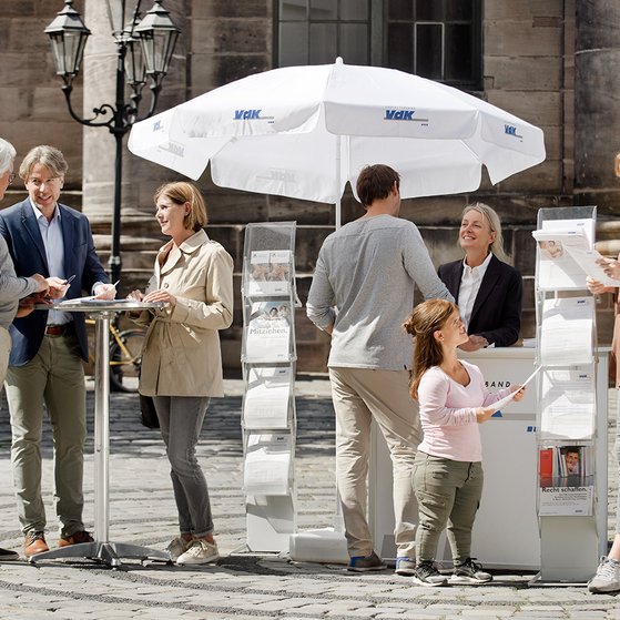 Ein VdK-Infostand in einer Fu&szlig;g&auml;ngerzone, eine Frau spricht mit einem Passanten, am Stehtisch daneben unterhalten sich Menschen. 