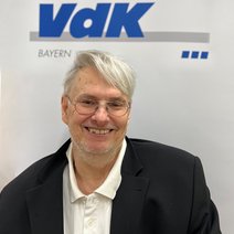 Auf dem Bild ist ein Mann mit kurzen grauen Haaren, Brille, wei&szlig;em Hemd und schwarzem Jackett zu sehen