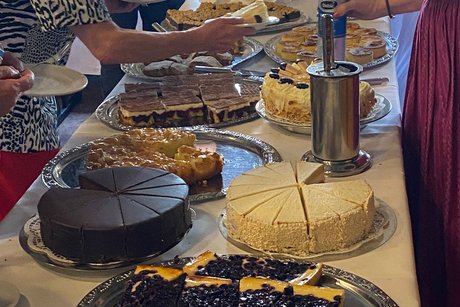 mehrere Kuchen stehen auf einem Tisch; einige G&auml;ste suchen sich ihre Kuchen aus.