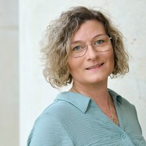 Auf dem Bild ist eine Frau mit kinnlangen gelockten blonden Haaren, Brille und schwarzem Oberteil zu sehen
