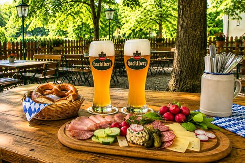 Brotzeit im Biergarten, auf dem Tisch stehen 2 Wei&szlig;biere und ein Brotzeitteller