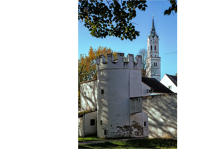 Das Bild zeigt einen Ausschnitt der Stadt Schrobenhausen bei strahlend blauen Himmel. Im Vordergrund ist die Stadtmauer abgebildet. Hinter der Stadtmauer kann man die Spitze eines Kirchturms sehen.