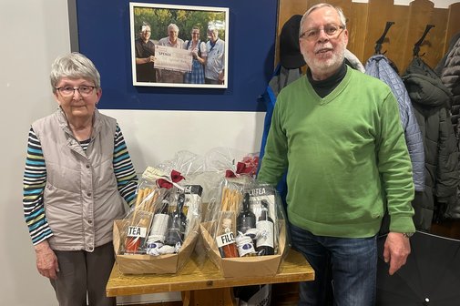 Margita Kleine-Nie&szlig;e und Peter Joist stehen links und rechts eines kleinen Tisches auf dem Ihre Pr&auml;sente in Form eines kleinen Geschenkkorbes stehen. Im Hintergrund eine Wand vom Sportrestaurant TV Ingolstadt Nord mit einem Teil der Garderobe.